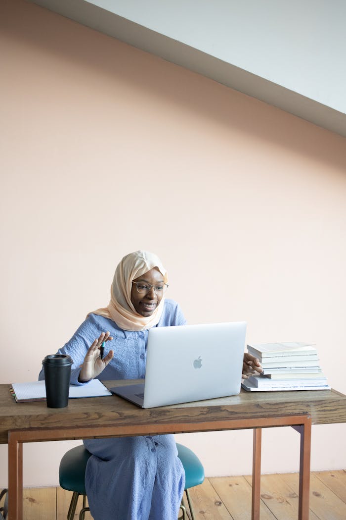 A woman in hijab waving at a laptop during a video call in a home office setting.