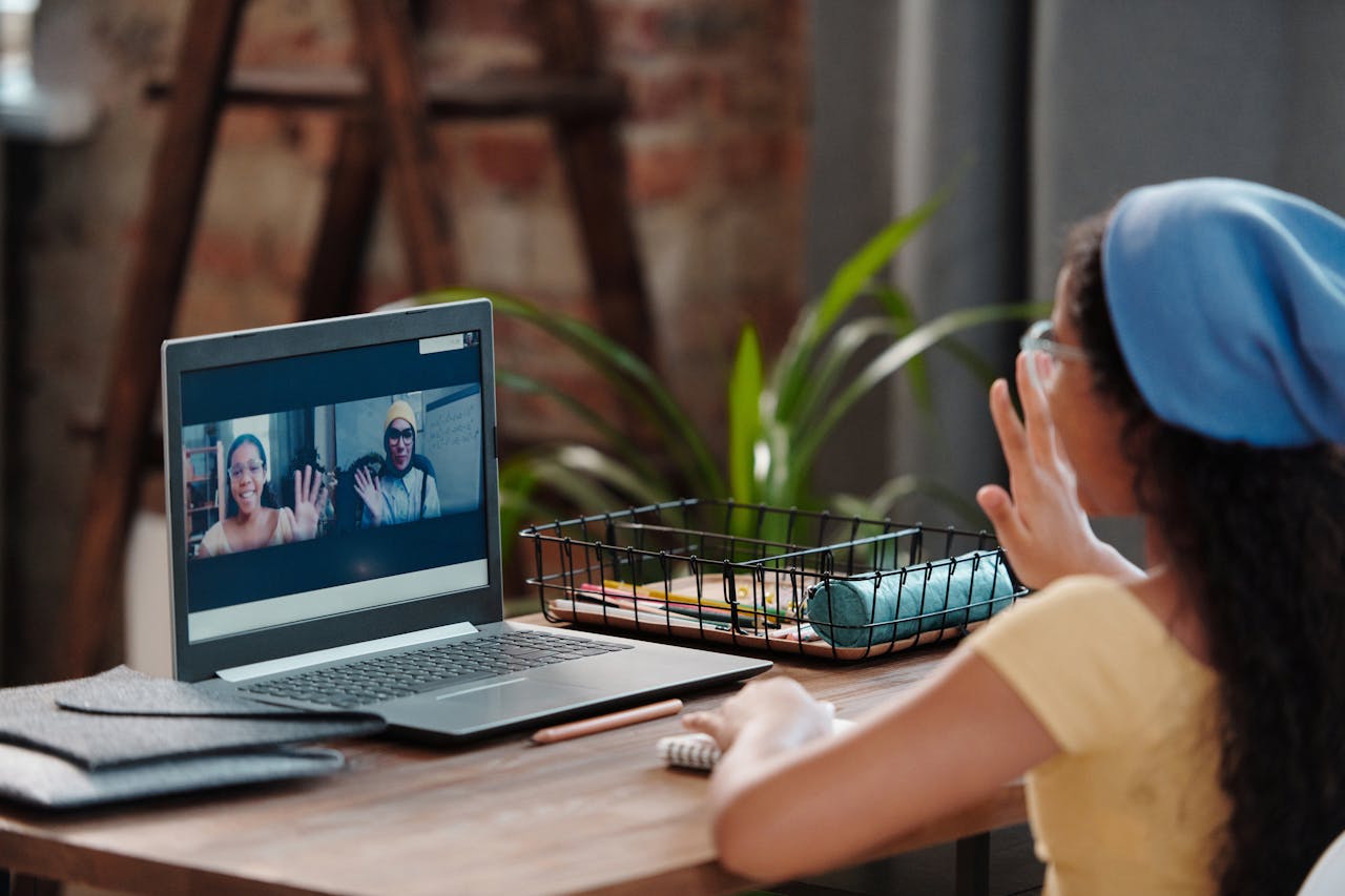 Young girl engaging in an online class from home, using a laptop and waving to her screen.